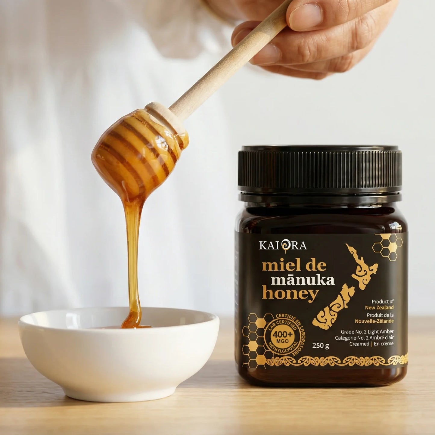 CUILLÈRE EN BOIS DE MIEL DU POT DE MIEL DE MANUKA DE 250 G DE MARQUE KAI ORA AVEC UN INDICE MGO 400+ QUI COULE VERS UN BOL